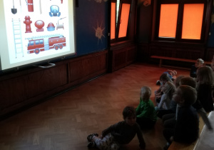 Na zdjęciu dzieci oglądające slajd na tablicy multimedialnej, na którym przedstawione są narzędzia wykorzystywane w pracy przez strażaka.